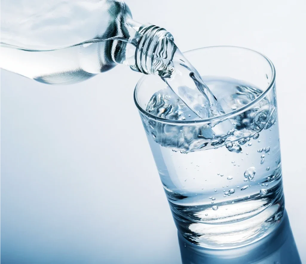 Close-up of a glass bottle of water pouring liquid into a glass sitting on a reflective surface.